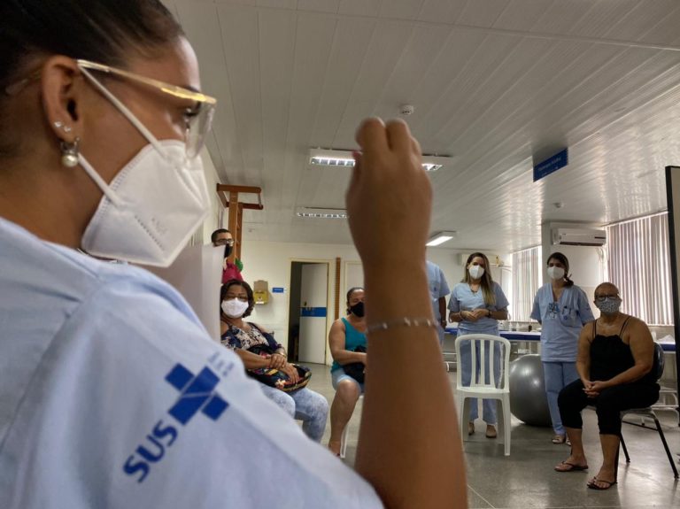 Em primeiro plano, uma mulher com uniforme azul com a marca do SUS na manga da camisa. Ela usa máscara e óculos de grau. Ao fundo aparece outras pessoas sentadas em cadeiras.