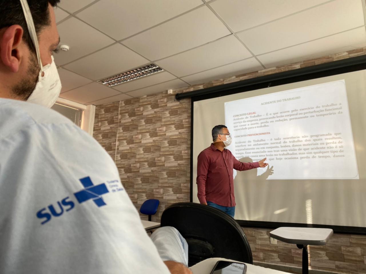 Em primeiro plano, a imagem mostra um colaborador do Multicentro Carlos Gomes sentado, de perfil, assistindo uma palestra sobre acidente de trabalho. Em segundo plano, à frente do projetor, o técnico de segurança do trabalho da unidade faz a apresentação da palestra.