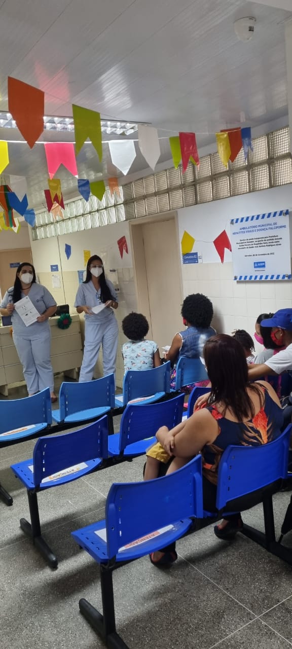 Duas colaboradoras do Multicentro Carlos Gomes estão com panfletos nas mãos, realizando uma palestra sobre doença falciforme numa sala de espera da unidade. Pacientes e acompanhantes aparecem de costas sentados nas cadeiras enquanto aguardam atendimento.