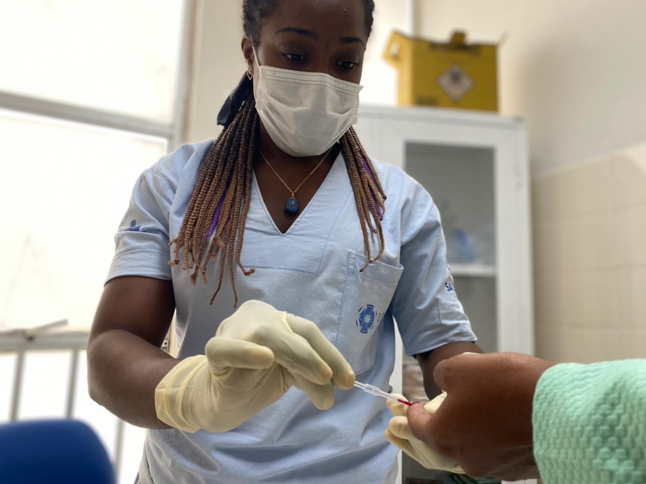 A técnica de enfermagem faz a coleta de amostra de sangue do dedo da paciente para realização do teste rápido. Ela usa máscara e luvas cirúrgicas.