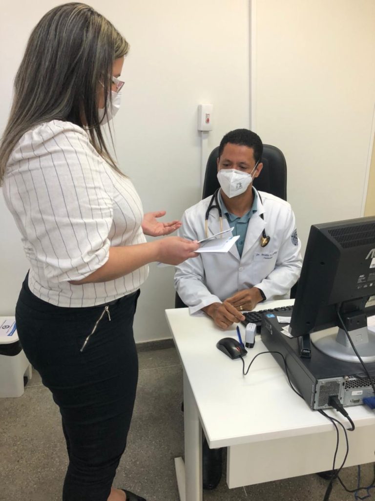 Uma mulher está de pé e aparece de perfil para câmara. Um homem com jaleco branco e máscara está sentado e apoia as mãos numa mesa. Na mesa tem um computador.