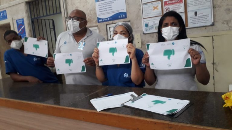 Três pessoas estão de pé atrás de um balcão de madeira. Elas seguram um cartaz educativo.