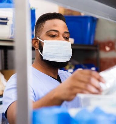 Em primeiro plano, aparece uma prateleira com insumos hospitalares com a imagem desfocada. Ao fundo, um homem com máscara e vestido com fardamento hospitalar arruma os materiais na prateleira.