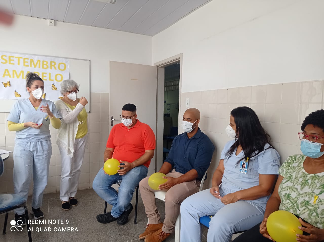 Duas pessoas estão de pé no fundo da sala e outras quatro estão sentadas uma ao lado das outras. Elas estão com balões amarelos nas mãos.