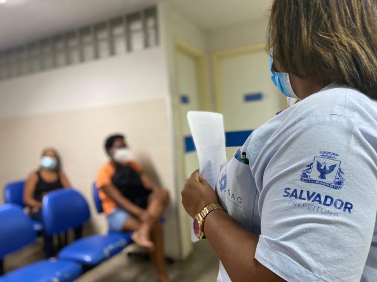 Em primeiro plano, está uma mulher de perfil com um uniforme azul com a logo da prefeitura de Salvador na manga da camisa e a marca do ISAC no lado esquerdo na altura do peito da camisa. Ao fundo, desfocado, duas mulheres estão sentadas em cadeiras azuis. Todos usam máscara.